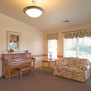 A cozy living room featuring a piano, floral sofa, and large windows letting in natural light.