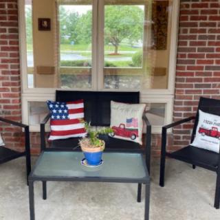 A cozy patio setup with chairs and a table, decorated with American flag and fire truck themed cushions, alongside a potted plant.