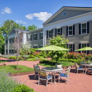 A well-maintained outdoor patio area with tables and chairs, surrounded by lush greenery in front of a large, colonial-style building.