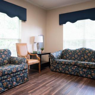 A cozy living room featuring floral patterned sofas, a wooden chair, window blinds, and a small lamp on a side table.
