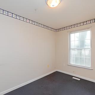 An empty room with a single window, neutral-colored walls, and dark carpeting.