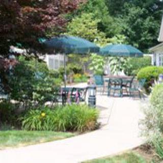 A sunny garden patio area with tables, chairs, and umbrellas surrounded by lush greenery.