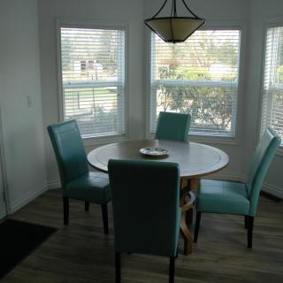 A cozy dining room with a round table and teal chairs, illuminated by natural light from the windows.