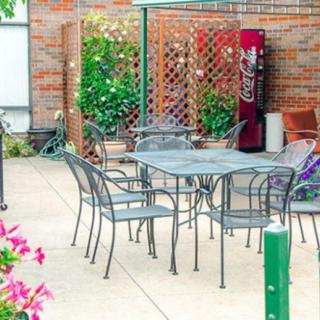 An outdoor patio area with metal table and chairs, surrounded by plants and a vending machine in the background.