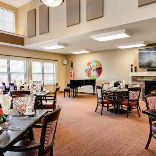 A well-lit dining area with tables, chairs, and a piano near large windows.