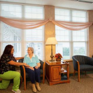 A caregiver kneels beside an elderly woman in a comfortable living room setting within a nursing home.