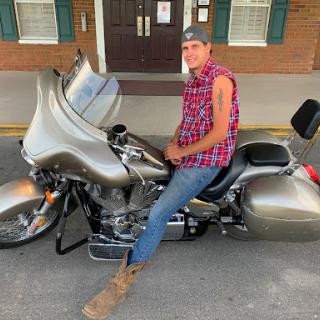 A man in a plaid shirt and jeans poses confidently on his motorcycle.