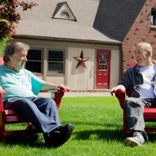 Two individuals sitting on red chairs, conversing on a grassy lawn in front of a house.
