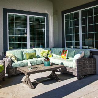 A cozy outdoor patio setup with a wicker sofa, colorful cushions, and a small coffee table in front of large windows.