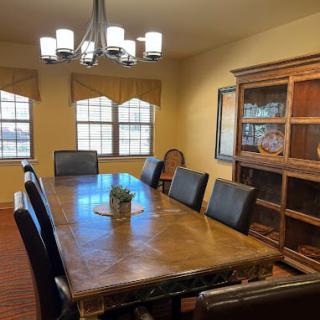 A spacious dining room with a large wooden table, surrounded by chairs and illuminated by a chandelier, featuring a cabinet and multiple windows.
