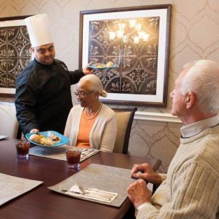 A chef serves food to elderly patrons at a dining table in a restaurant.