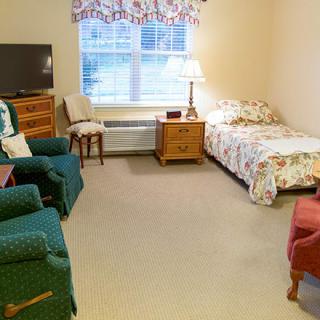 A cozy bedroom with a single bed, armchairs, a TV, and a window providing natural light.