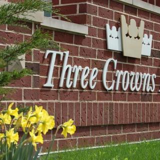 A brick wall featuring a sign that reads 'Three Crowns', with yellow daffodils in the foreground.