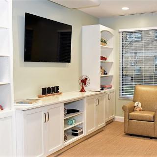 A cozy living room featuring a television mounted on the wall, surrounded by white shelves, with an armchair and window completing the setting.