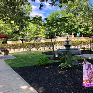 A serene garden with a decorative fountain, surrounded by lush greenery and a sunlit path.