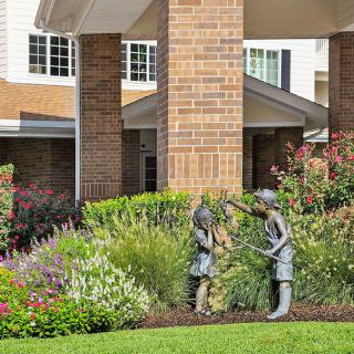 A garden scene with a statue of children surrounded by lush greenery in front of a brick building.