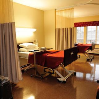 A view of a patient room in a hospital, featuring two beds with red covers and curtains for privacy.