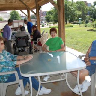A group of people sitting around a glass table under a gazebo in a park.