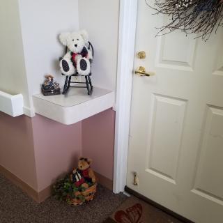 A cozy corner featuring a teddy bear on a rocking chair, a miniature car, and a basket of greenery beside a door.