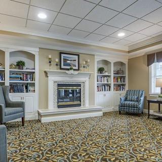 A cozy living room with a fireplace, surrounded by bookshelves and armchairs, featuring a patterned carpet and ceiling lights.