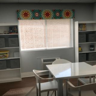 A cozy dining area with a white table and chairs, flanked by bookshelves and a window adorned with a colorful valance.