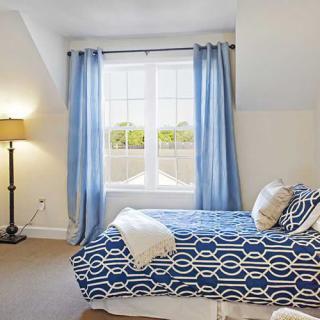 A cozy bedroom featuring a neatly made bed with a blue patterned duvet, soft blue curtains framing a bright window, and a stylish floor lamp illuminating the space.