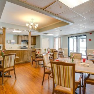 An empty dining room with wooden floors, several tables and chairs, and a kitchen area in the background.