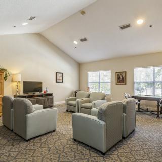 A cozy living room with armchairs arranged around a television, featuring large windows and carpeted flooring.