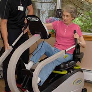 A senior individual using exercise equipment with the assistance of a trainer.