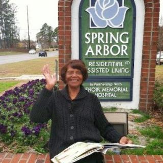 A woman happily waving while sitting in front of the Spring Arbor assisted living facility.