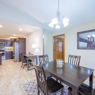 A modern dining room with a dark wooden table and chairs, adjacent to an open kitchen area, under a stylish light fixture.
