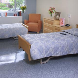 A bright and tidy hospital room featuring two patient beds, a cozy armchair, and decorative flowers by the window.