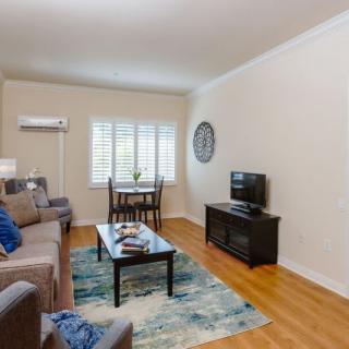 A cozy living room featuring a sofa, a television on a stand, a coffee table with decor items, and a small dining area near the window.