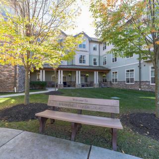 A peaceful outdoor area with a wooden bench in front of a residential building surrounded by trees and a well-maintained lawn.