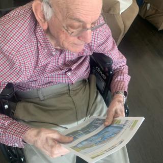 An elderly man seated in a wheelchair, reading a newspaper indoors.