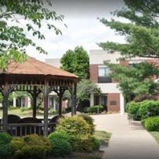 A cozy gazebo near a brick building surrounded by greenery and a paved pathway.