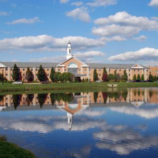 A large building with a spire reflecting in a calm lake under a partly cloudy sky.