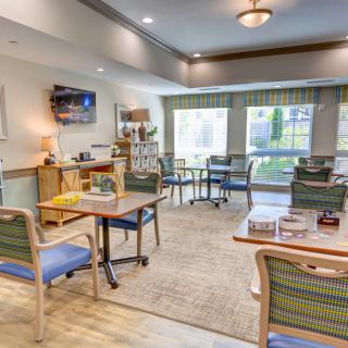 A cozy interior room with chairs, tables, and a television set, featuring large windows for natural light.