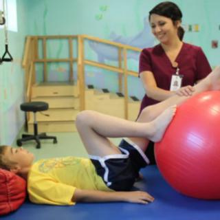 A therapist assists a child with leg exercises using a fitness ball during a physical therapy session.