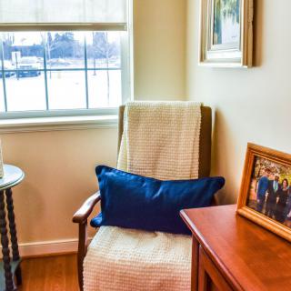 A cozy corner of a living room featuring an armchair with a blue pillow, a window with blinds, and a wooden table displaying family photos.