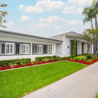 A single-story house with a well-maintained lawn, flower beds, and palm trees under a partly cloudy sky.