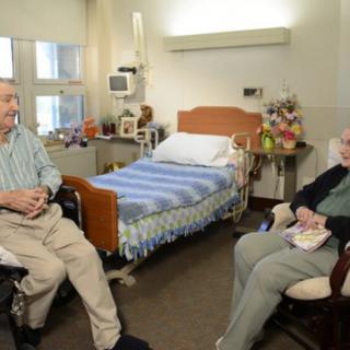 Two elderly individuals sitting in a hospital room, engaged in conversation.