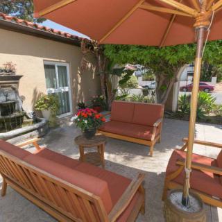 A cozy patio setup featuring wooden outdoor furniture with red cushions and a large umbrella amidst garden greenery.