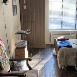 A cozy bedroom with natural sunlight streaming through the window, featuring a bed, wardrobe, and chair.