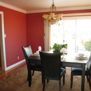 A cozy dining room with red walls, featuring a table and chairs, illuminated by a chandelier and natural daylight through the window.