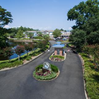 A scenic entrance with a roundabout and landscaped gardens, surrounded by trees and a pond, leading to buildings.