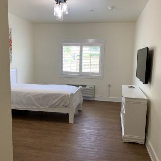 A minimalist bedroom featuring a bed, dresser, and wall-mounted TV, with natural light streaming through the window.