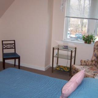 A cozy bedroom with a blue bedspread, a chair, and a window letting in natural light.