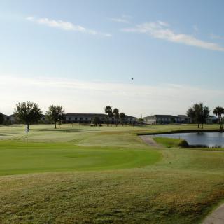 A scenic view of a golf course with lush green grass, trees, and a pond under a clear morning sky.