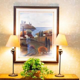 A cozy interior setting featuring a framed landscape painting above a console table, flanked by two elegant table lamps and adorned with a lush green plant.